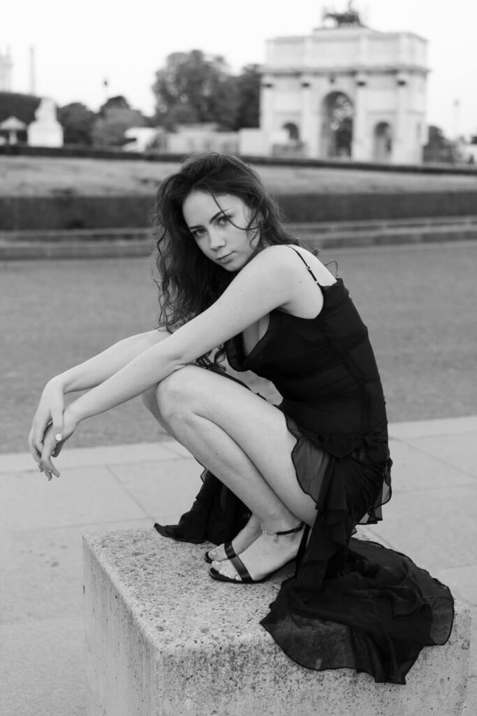 a portrait photography of a young women, wearing a cocktail dress is crouching on a stone on the square in Paris