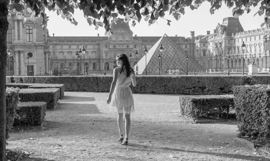 A portrait of a girl walking in the park with her back turned in front of the Louvre Museum in Paris