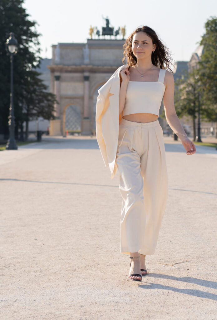 A young woman dressed in a cream trouser suit is walking in the park in Paris with the Triumph Arch in the background