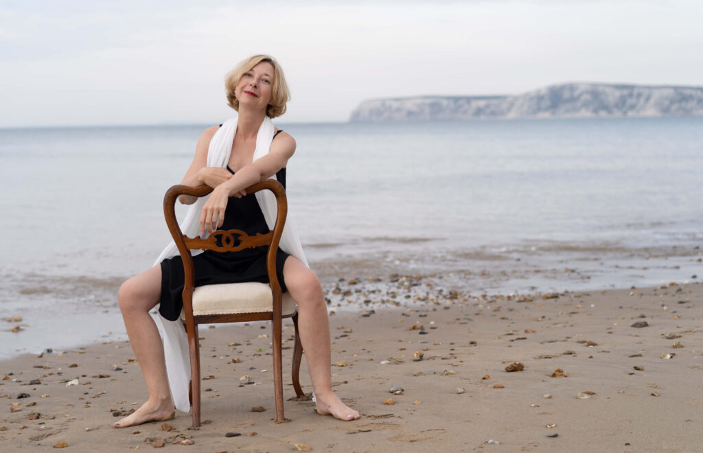 A portrait of a beautiful women sitting on a chair by the sea. She is dressed in a black dress and wearing a white scarf.